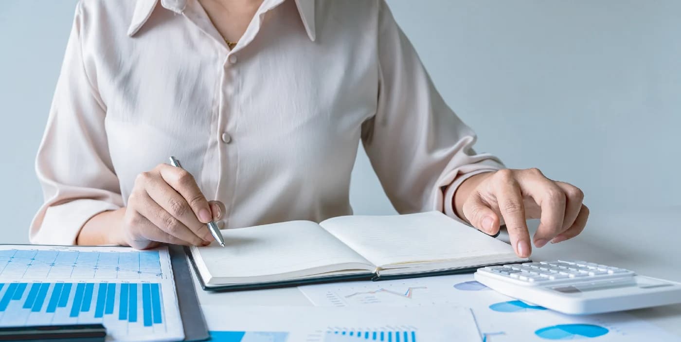 Hand writing in a notebook beside printed financial charts and a calculator