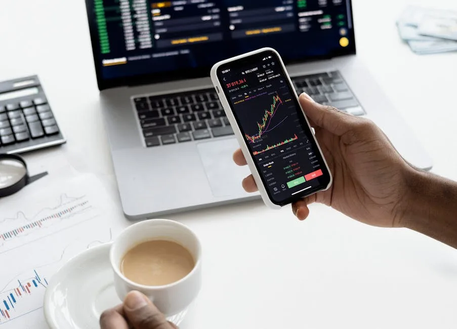 Laptop and phone showing financial charts beside a coffee on a desk